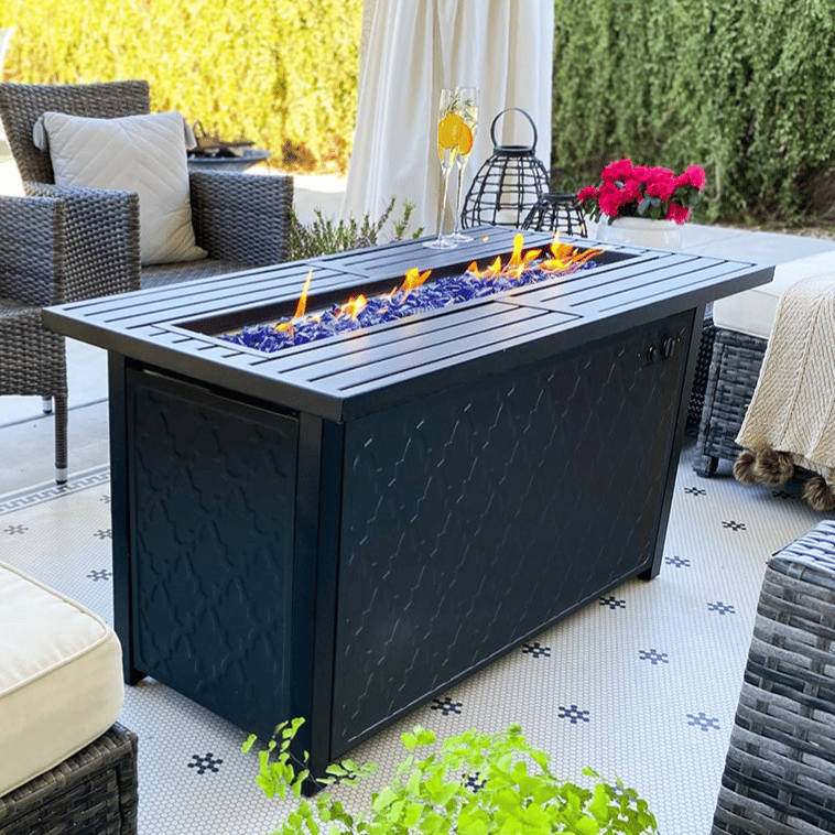 A stylish outdoor patio set with wicker chairs, a cushioned seat, and a sleek rectangular fire pit table featuring blue glass stones. Small potted plants and decorative items add to the inviting atmosphere.