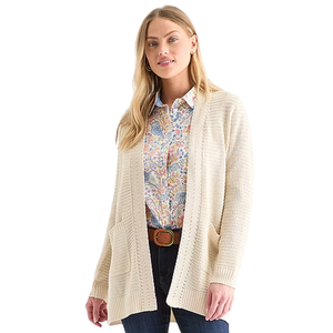 A woman wears a beige knitted cardigan over a colorful floral shirt, paired with dark jeans and a brown belt.