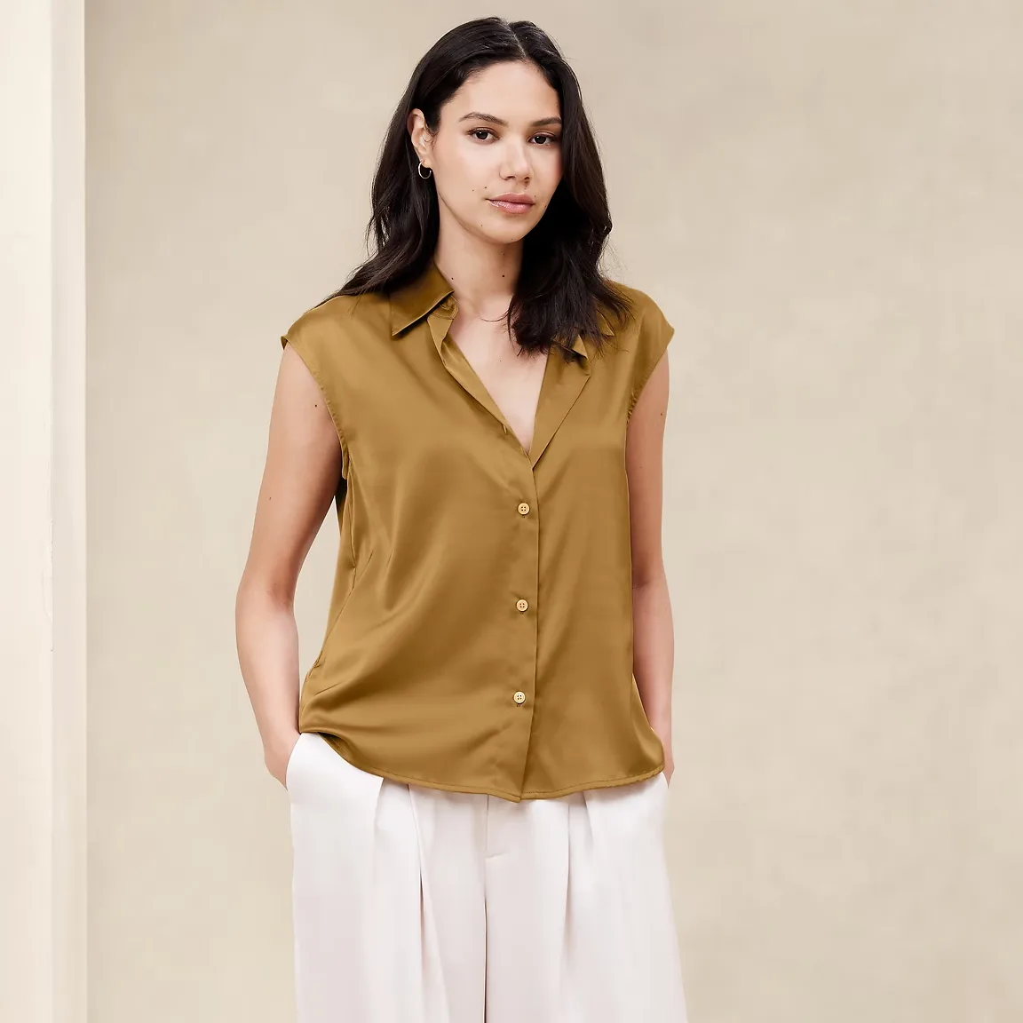 A woman is wearing a sleeveless olive green button-up blouse and white pants.