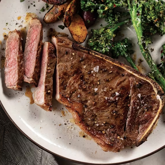 A cooked T-bone steak is on a plate, sliced and seasoned, accompanied by roasted potatoes and broccolini.