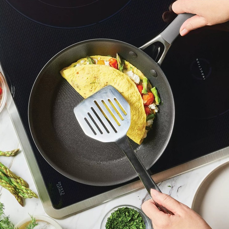 A person is using a spatula to lift an omelet filled with asparagus, tomatoes, and cheese in a non-stick frying pan on an induction cooktop. Fresh herbs and asparagus spears are visible nearby.