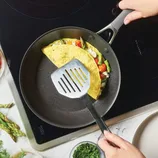 A person is using a spatula to lift an omelet filled with asparagus, tomatoes, and cheese in a non-stick frying pan on an induction cooktop. Fresh herbs and asparagus spears are visible nearby.