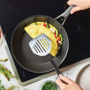A person is using a spatula to lift an omelet filled with asparagus, tomatoes, and cheese in a non-stick frying pan on an induction cooktop. Fresh herbs and asparagus spears are visible nearby.