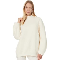 A woman wears a white, oversized, ribbed-knit sweater from Free People. The sweater features a high neckline and long sleeves, providing a cozy and relaxed look.
