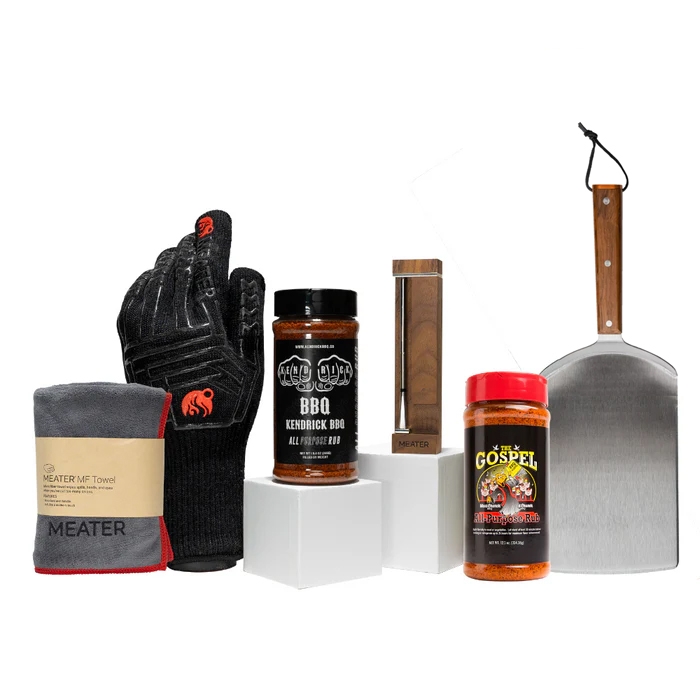 BBQ tool set featuring a black heat-resistant glove, a microfiber towel, two jars of BBQ seasoning, a MEATER wireless meat thermometer, and a pizza peel with a wooden handle.
