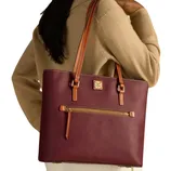 A woman is carrying a plum-colored leather tote with tan straps and a front zipper pocket, featuring a logo plaque. She's wearing a beige sweater and white pants.