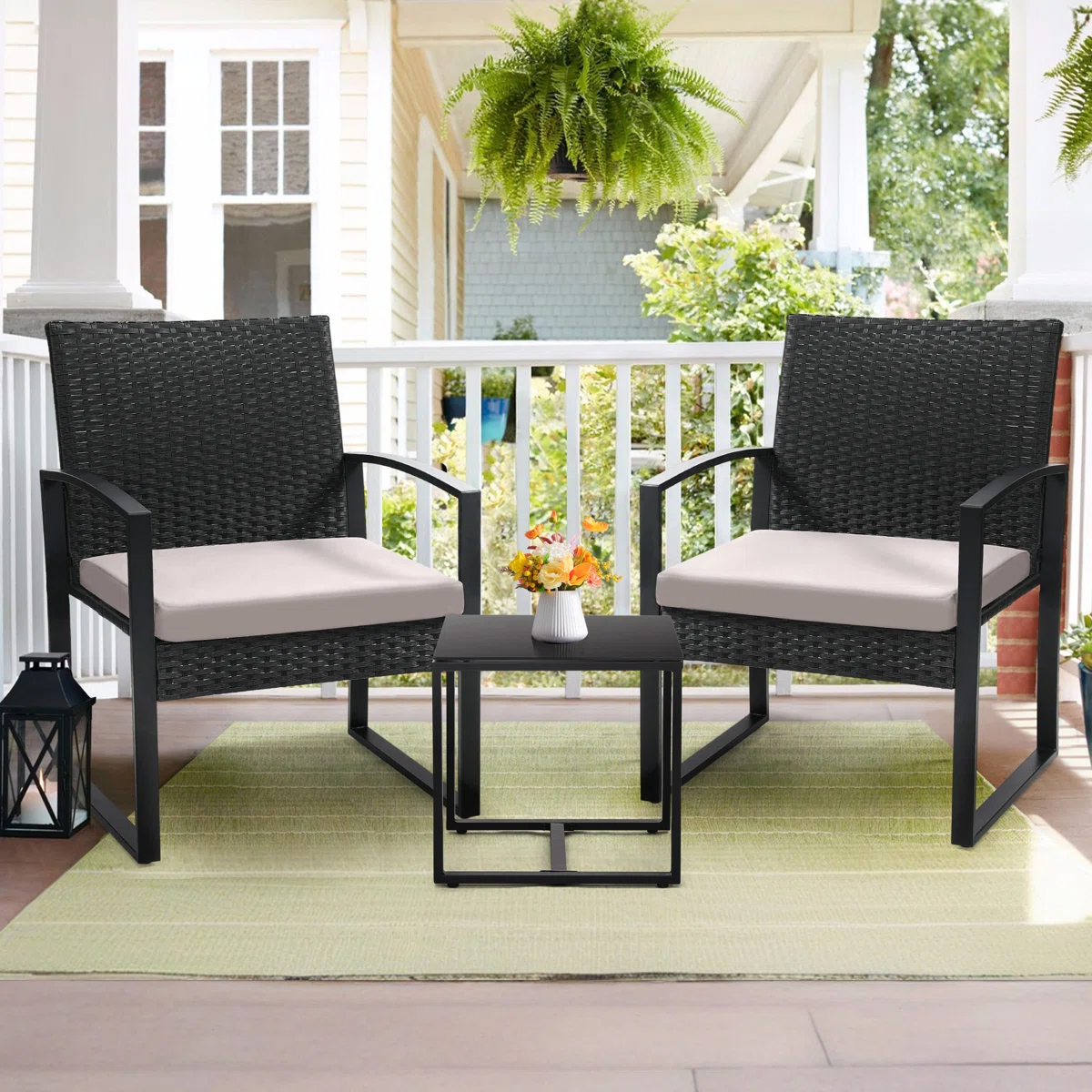 Two black wicker chairs with beige cushions are on a porch, accompanied by a small black table with a white vase containing flowers. A green rug is beneath, and there's a hanging fern and a lantern nearby.