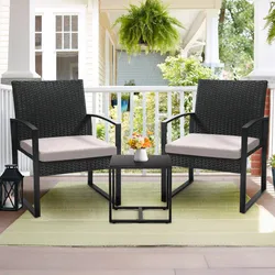 Two black wicker chairs with beige cushions are on a porch, accompanied by a small black table with a white vase containing flowers. A green rug is beneath, and there's a hanging fern and a lantern nearby.