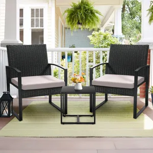 Two black wicker chairs with beige cushions are on a porch, accompanied by a small black table with a white vase containing flowers. A green rug is beneath, and there's a hanging fern and a lantern nearby.