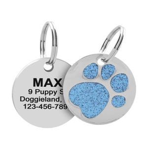 A pet ID tag with a blue glitter paw print design on one side and personalized text on the other, including the pet's name \“MAX,\“ address, and phone number.