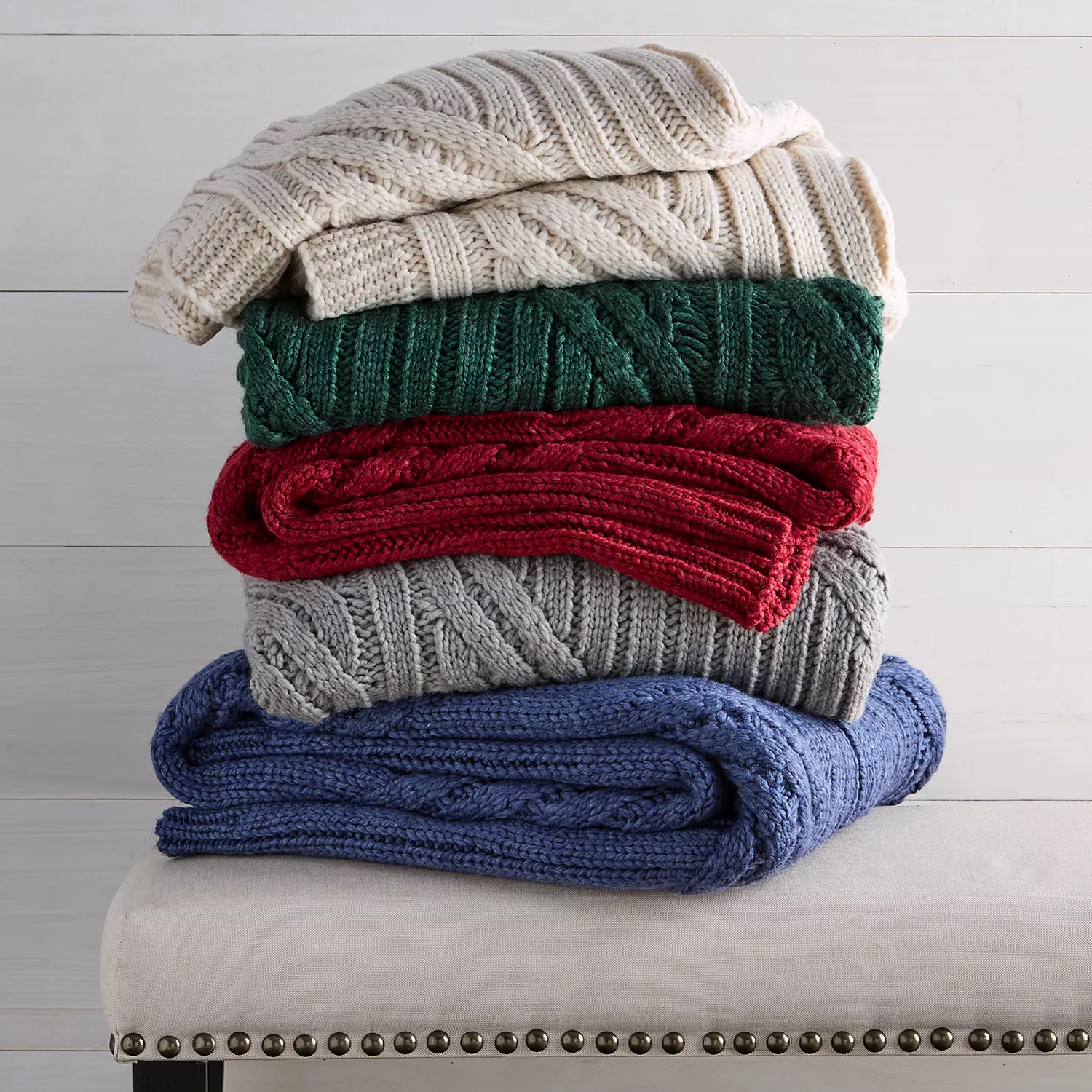 A stack of five cable-knit blankets in cream, green, red, gray, and blue is neatly arranged on a bench with nailhead trim.