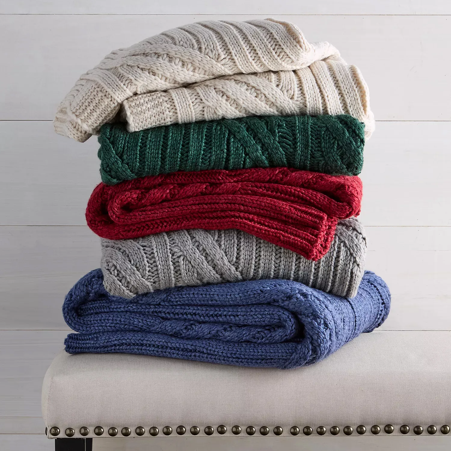 A stack of five cable-knit blankets in cream, green, red, gray, and blue is neatly arranged on a bench with nailhead trim.