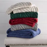 A stack of five cable-knit blankets in cream, green, red, gray, and blue is neatly arranged on a bench with nailhead trim.