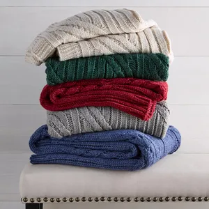 A stack of five cable-knit blankets in cream, green, red, gray, and blue is neatly arranged on a bench with nailhead trim.