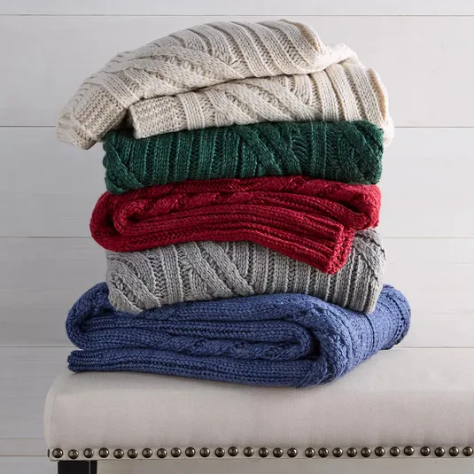 A stack of five cable-knit blankets in cream, green, red, gray, and blue is neatly arranged on a bench with nailhead trim.