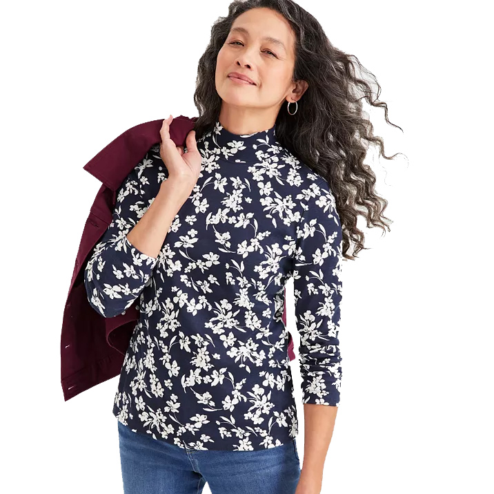 A woman is wearing a navy blue cotton-blend long-sleeve top with a white floral pattern and a mock neck. She is holding a burgundy jacket over her shoulder and is paired with blue jeans.