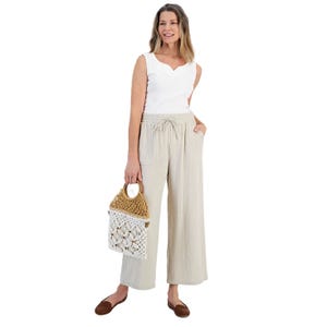 A woman is wearing a white tank top and light beige wide-leg pants, holding a crochet bag with a wooden handle. She is also wearing brown flats.