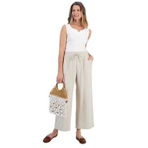 A woman is wearing a white tank top and light beige wide-leg pants, holding a crochet bag with a wooden handle. She is also wearing brown flats.