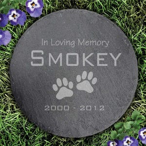 Round slate garden stone with \“In Loving Memory, Smokey\“ and paw prints etched; years 2000-2012, surrounded by grass and purple flowers.