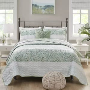 A green and white seersucker quilt set with a floral pattern includes a quilt and matching pillow shams. The bed features a decorative green cushion, adjacent white nightstands with lamps, and a cozy knitted pouf at the foot.