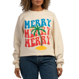 A woman is wearing a beige sweatshirt with colorful, bold text that says \“MERRY MERRY MERRY\“ around a palm tree. She pairs it with blue jeans.