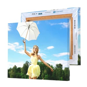 A canvas print measuring 11\“ x 14\“ shows a woman in a yellow dress holding a white umbrella, set against a bright blue sky and lush green trees, ready to be hung as wall art. The frame is wooden with canvas stretching details visible.