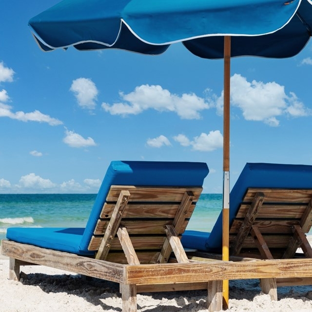 Two wooden beach loungers with blue cushions sit under a blue umbrella on a sandy beach, facing the ocean.