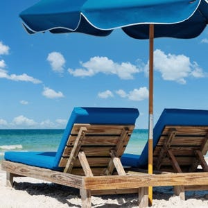 Two wooden beach loungers with blue cushions sit under a blue umbrella on a sandy beach, facing the ocean.
