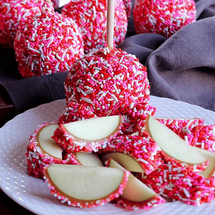 Apples coated in pink and white sprinkles, skewered on sticks, with some sliced pieces displayed on a white plate.
