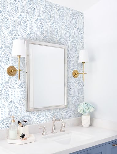 A bathroom vanity setup with a blue and white patterned wallpaper, featuring a rectangular mirror, brass wall-mounted lights with white shades, a white countertop with a chrome faucet, and blue cabinetry. A vase with white flowers and toiletries adorn the counter.