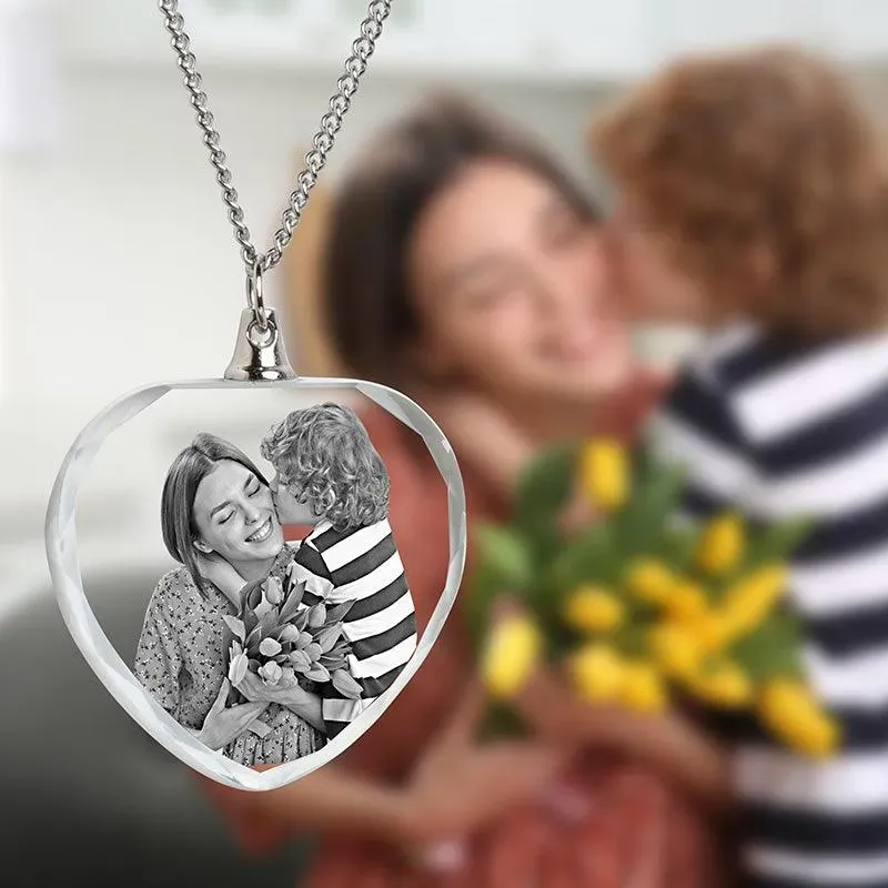 A heart-shaped crystal necklace features a black-and-white image of a smiling woman receiving a kiss and flowers from a child, with a blurred background of the original scene.