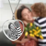 A heart-shaped crystal necklace features a black-and-white image of a smiling woman receiving a kiss and flowers from a child, with a blurred background of the original scene.