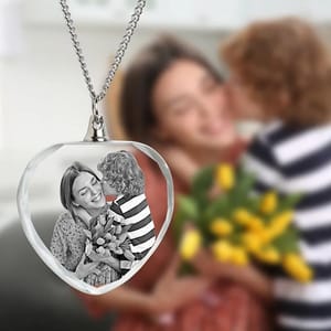 A heart-shaped crystal necklace features a black-and-white image of a smiling woman receiving a kiss and flowers from a child, with a blurred background of the original scene.