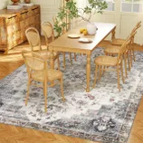 A dining room setup features a wooden table with six cane-backed chairs on a decorative, floral-patterned rug. A rustic dresser and a plant in a vase add decorative elements.