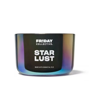 A cylindrical, iridescent glass candle labeled \“FRIDAY COLLECTIVE. STAR LUST\“ indicating it is made with essential oils.
