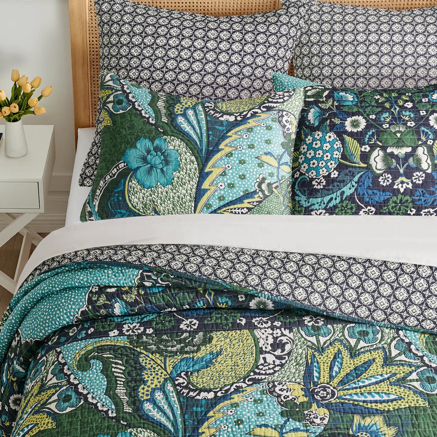 A bedding set featuring a quilt, pillowcases, and shams in vibrant floral and geometric prints with shades of green, blue, and black. A small vase with yellow flowers sits on a white side table nearby.