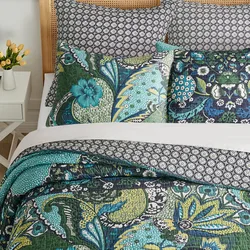 A bedding set featuring a quilt, pillowcases, and shams in vibrant floral and geometric prints with shades of green, blue, and black. A small vase with yellow flowers sits on a white side table nearby.