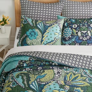 A bedding set featuring a quilt, pillowcases, and shams in vibrant floral and geometric prints with shades of green, blue, and black. A small vase with yellow flowers sits on a white side table nearby.