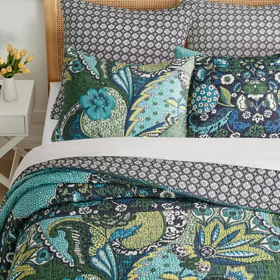A bedding set featuring a quilt, pillowcases, and shams in vibrant floral and geometric prints with shades of green, blue, and black. A small vase with yellow flowers sits on a white side table nearby.