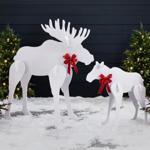 White silhouette lawn figures of a moose and a calf with red bows, set on artificial snow between two lit Christmas trees.