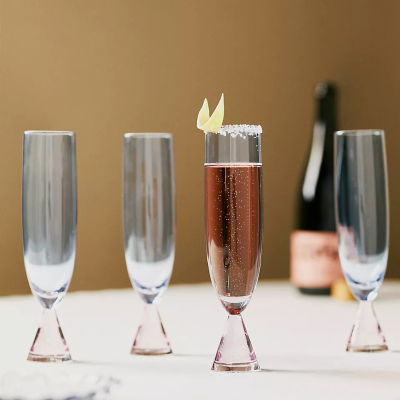 Four clear champagne flutes, with one filled with a sparkling pink liquid, garnished with a lemon twist and sugar rim. A bottle is blurred in the background.