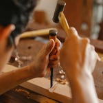A person is using a woodworking chisel and a mallet on a wooden piece clamped to a workbench.