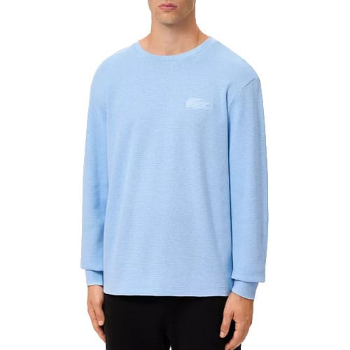 A man is wearing a light blue long-sleeve sweatshirt with a subtle logo on the chest, paired with black pants.