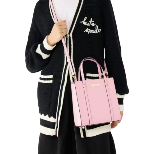 A pink Kate Spade Kenzie Small Tote is shown, featuring two top handles, a detachable shoulder strap, and gold-tone hardware. The model is wearing a black cardigan with white trim and a logo, paired with a black skirt.