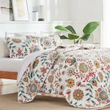 A Queen Floral Quilt Set featuring a vibrant floral pattern in red, green, yellow, and blue on a white background. Includes a quilt and matching pillow shams, adding a colorful and cozy touch to any bedroom.