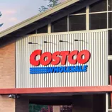Costco Wholesale storefront featuring its recognizable red and blue logo. The building has large windows and a modern architectural design with a backdrop of trees.