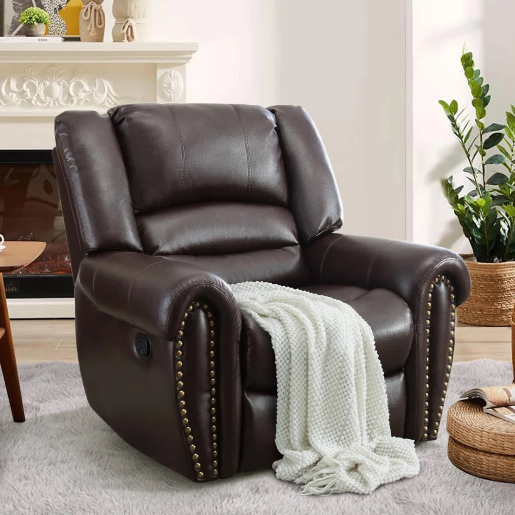 A dark brown leather recliner with stud accents is placed on a light rug, draped with a white knitted blanket. It sits next to a plant and a round wicker side table with magazines, in a cozy room setting.