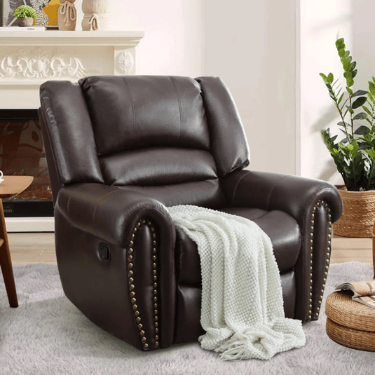 A dark brown leather recliner with stud accents is placed on a light rug, draped with a white knitted blanket. It sits next to a plant and a round wicker side table with magazines, in a cozy room setting.
