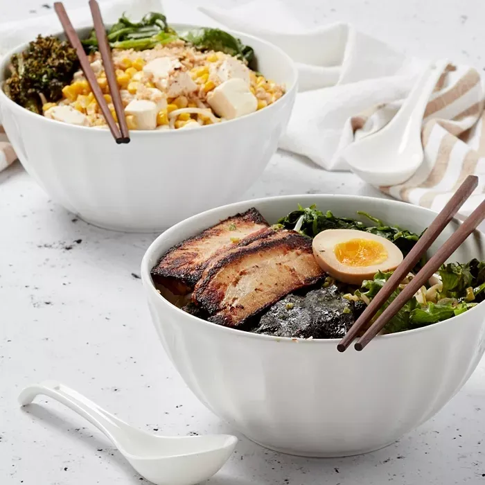Two bowls of Asian-inspired dishes: one with ramen, pork, seaweed, and egg and the other with rice, tofu, corn, and greens.