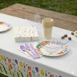A table setting includes floral-patterned paper plates, napkins, and a tablecloth, alongside translucent plastic bowls, lavender plastic cutlery, and stacked beige cups, set outdoors.